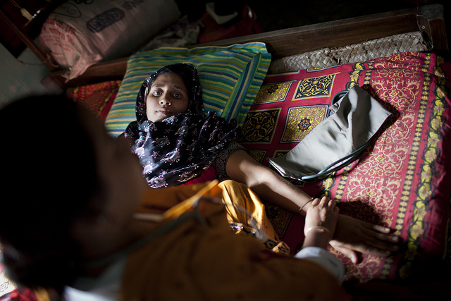 A pregnant woman lays on a bed of blankets with her arm extended, as another woman with medical tools touches her arm.