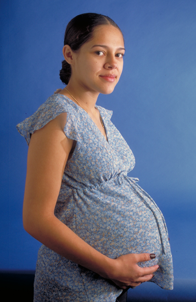 A pregnant woman in a blue dress holds her stomach and gazes at the camera.