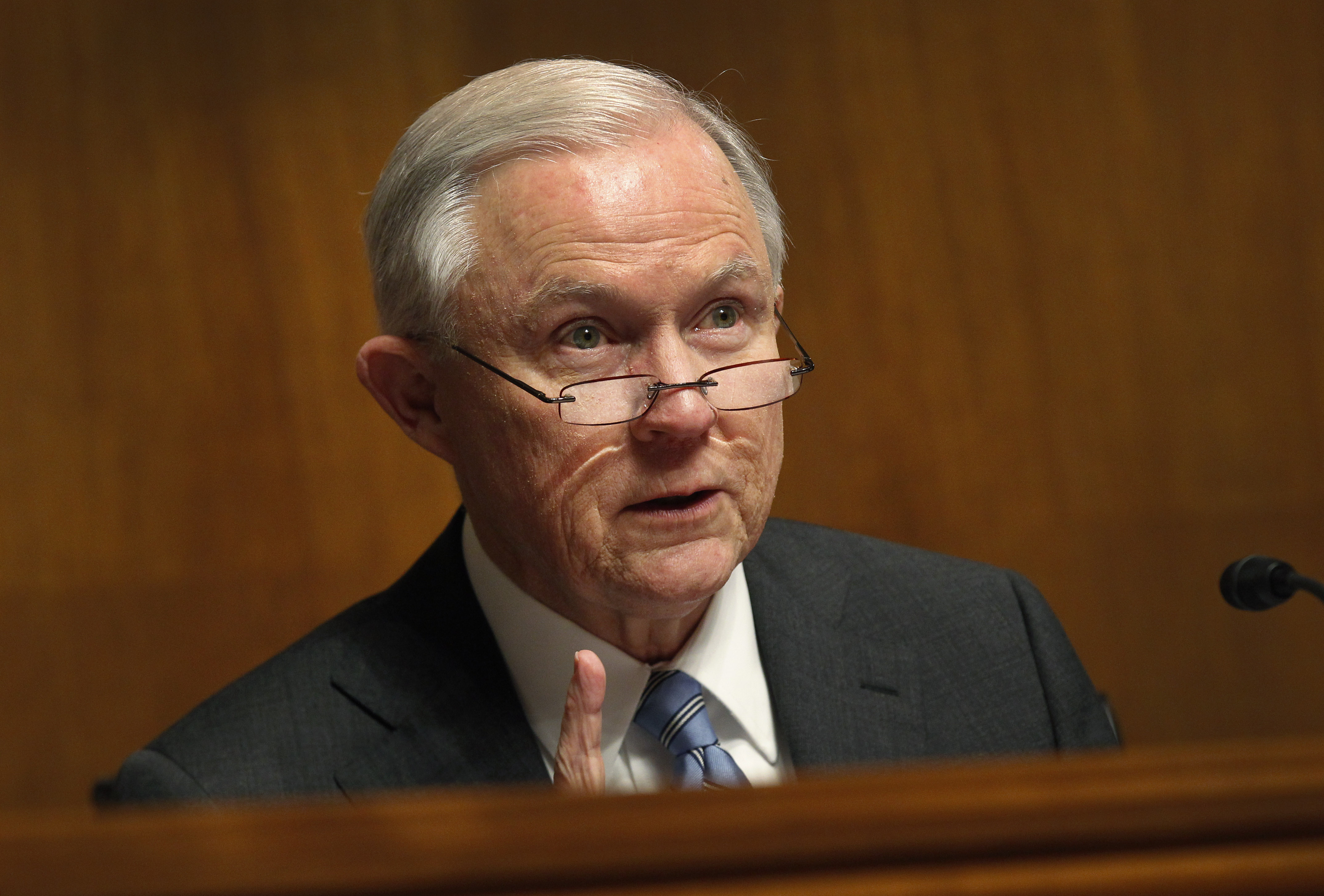 Jeff Sessions, a white male, wear a pair of glasses. He sits behind a desk and points a finger, about to make a statement.