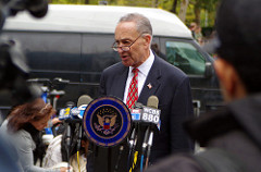 Senator Schumer stands behind a podium, surrounded by press.