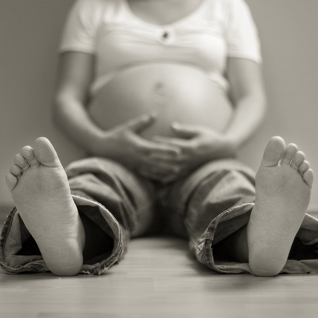 Grayscale image of a pregnant woman, with exposed belly, sitting with her bare feet in focus in front of her. Her face is not shown, and the picture displays only her body from the shoulders down.