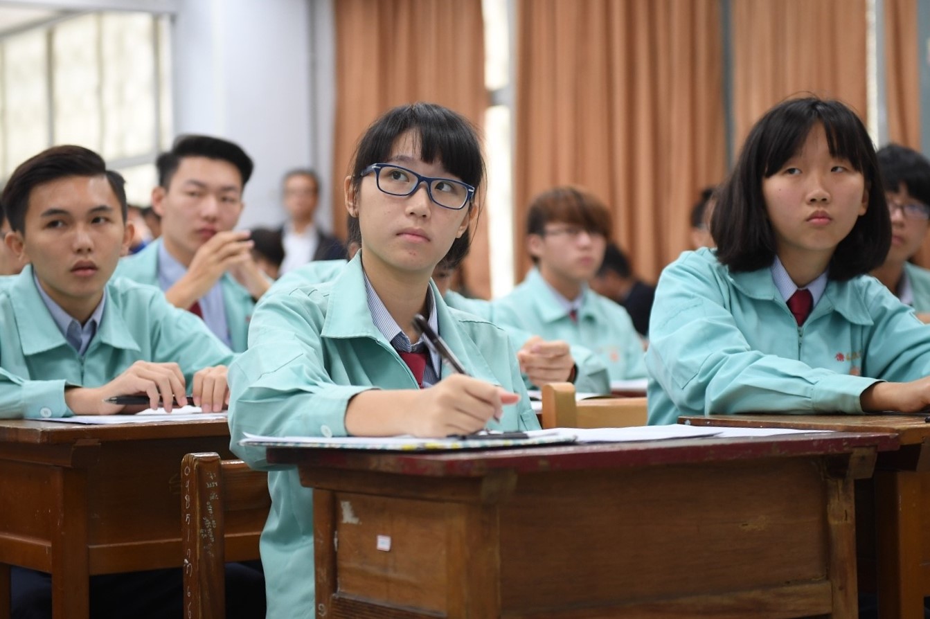 Rows of students wearing blue school uniforms sit at desks and take notes.