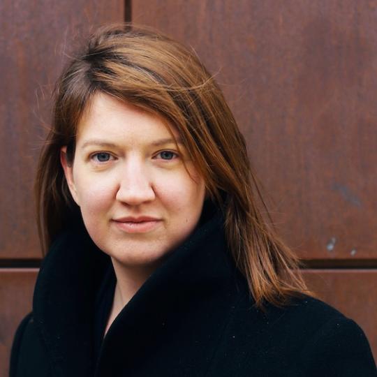 a  woman with brown hair in front of a brown wall wearing a black coat
