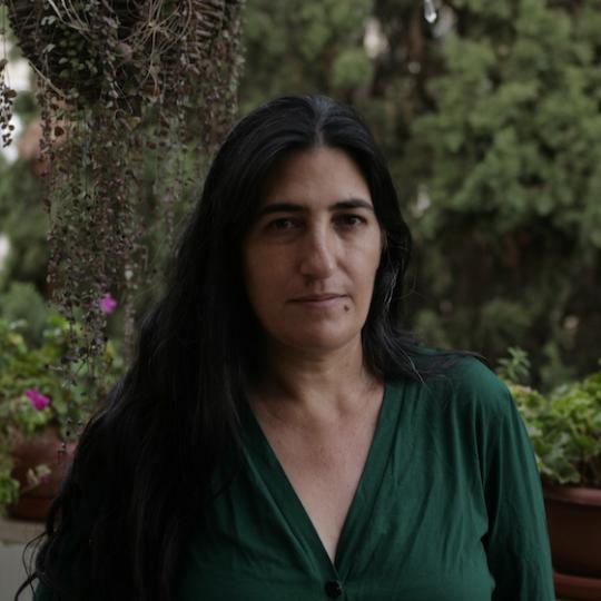 a woman with long black hair stands in front of hanging foliage