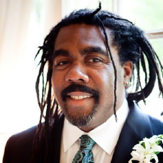 a Black man with grey dreadlocks in a suit with a flower