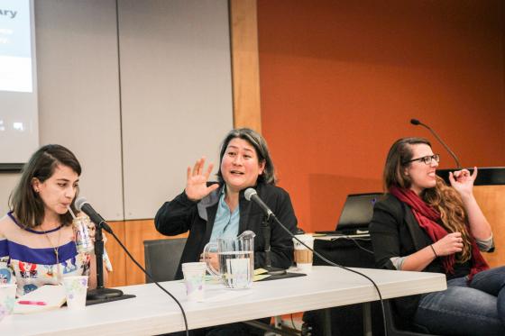 Karen Nakamura, seated behind a table, gestures as she speaks. On the left, Sara Acevedo sips a beverage. On the right, the ASL interpreter.