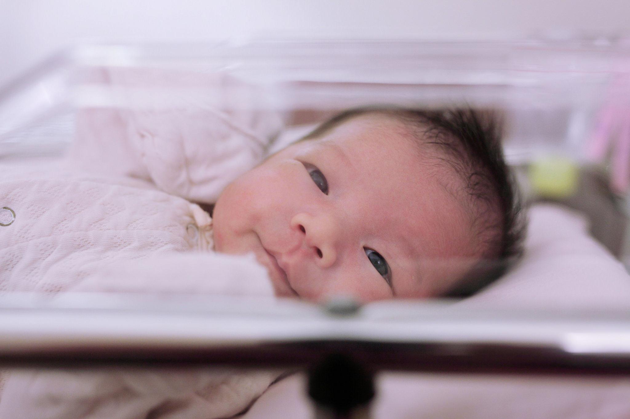 Close up photo of a baby through a clear hospital bed. The baby looks happily toward the camera.