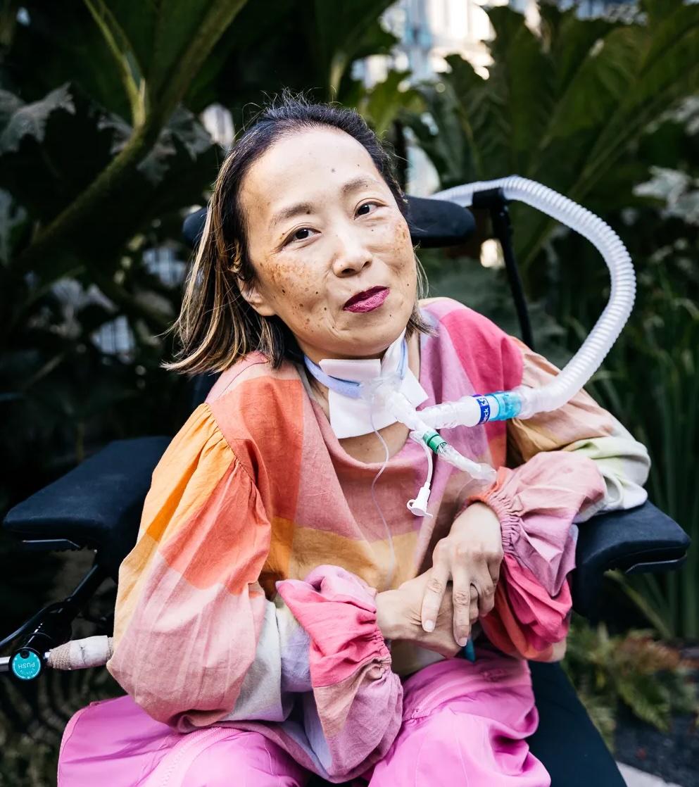 a photo of Alice Wong in an orange and pink outfit, seated in a powered wheelchair 