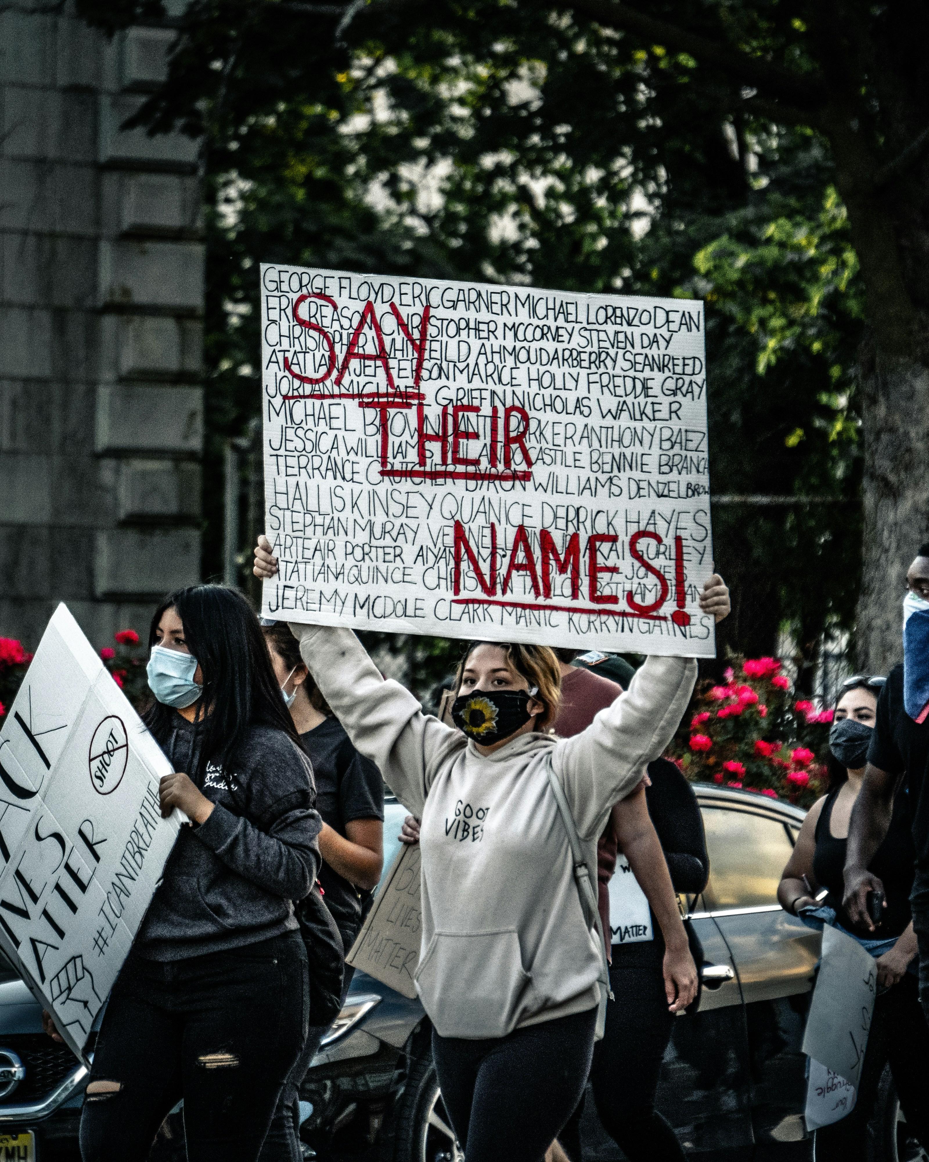 a woman holds up a sign at a protest that states "Say Their Names" with victims of police brutality listed on the poster