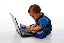 A young baby is fully dressed and sits in front of a laptop, interacting with the keyboard.