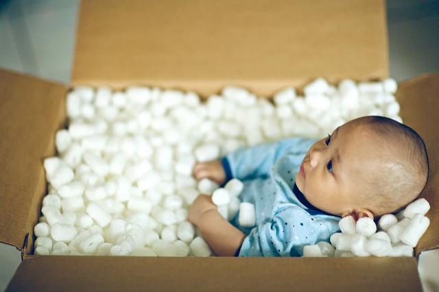 A baby stiffly lays in a cardboard box, surrounded by package bubble wrap.