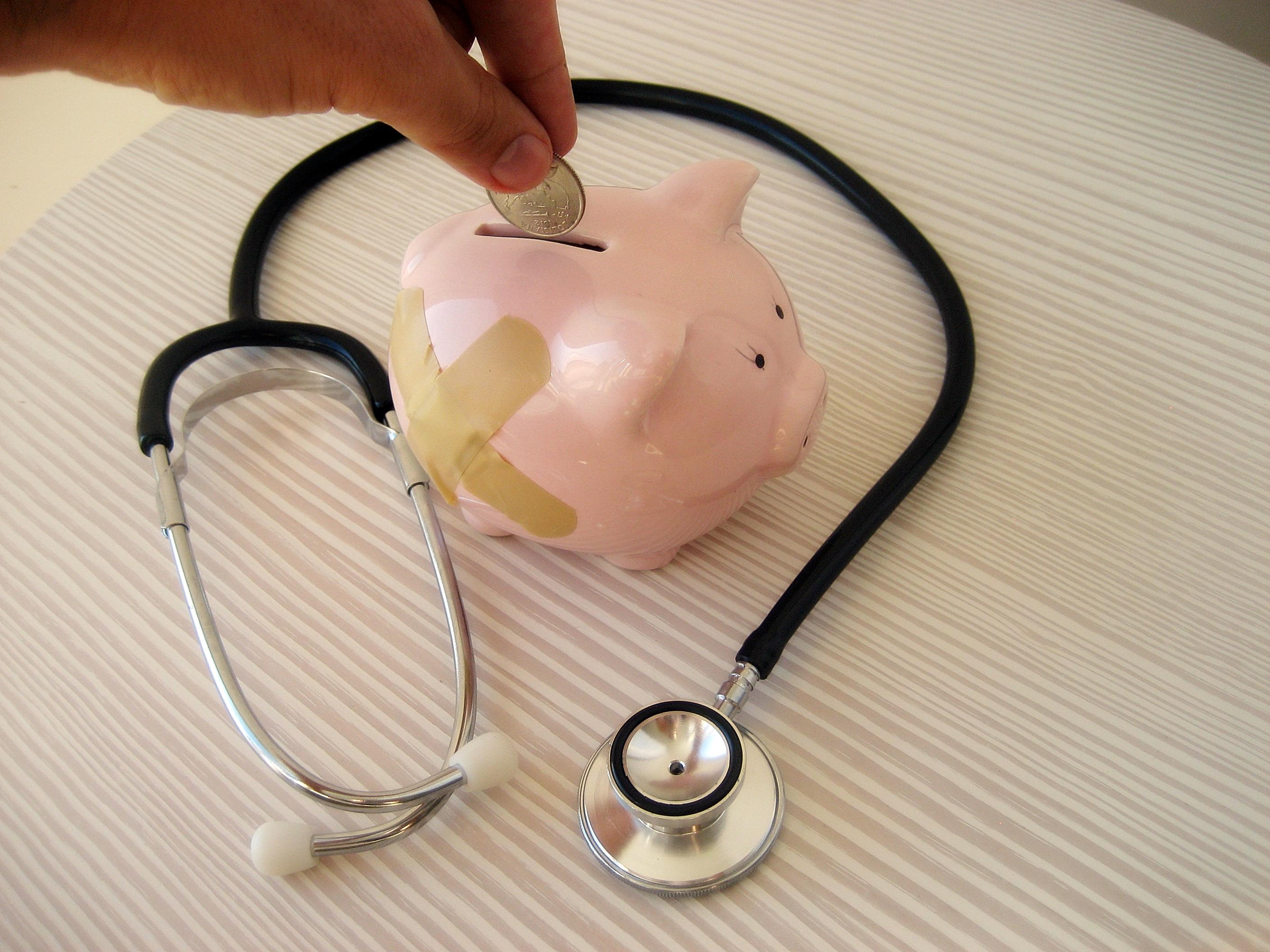 Someone puts a coin into a piggy bank that has bandages on its side. A doctor's stethoscope surrounds the piggy bank.