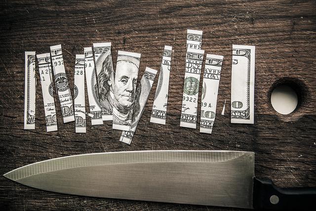 A one-hundred dollar bill is cut into pieces on  a cutting board. Below the bill is a kitchen knife.