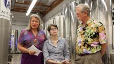 Screenshot from promotional video, which shows three white adults surrounded by large metal cylinders along the wall, known as the outpatient storage room. One of whom is the director of Alcor, another who is a young adult customer, and the last is a white male doctor dressed in a tropical shirt. 