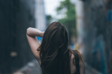 A woman is turned away from the camera walking through an alley between two cement buildings. Her left arm smoothens her hair.