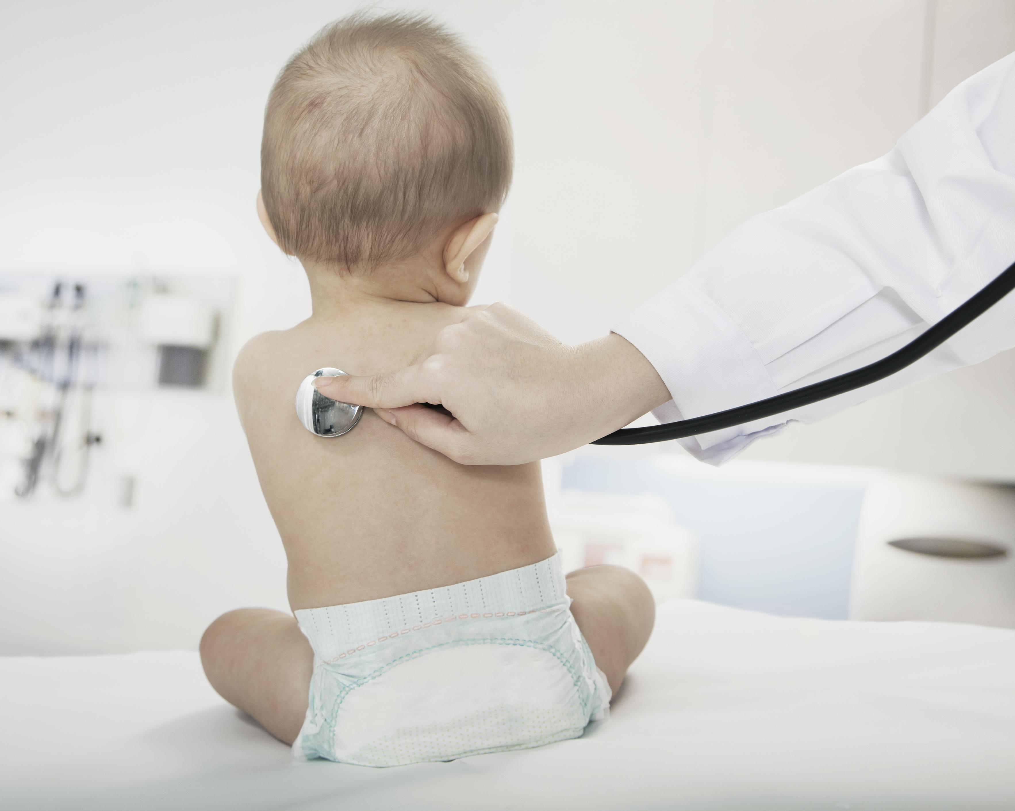 a blond a pale skin sits in a diaper on the bed in a doctors office. A doctor with only his arm in frame puts a stethoscope to their back.