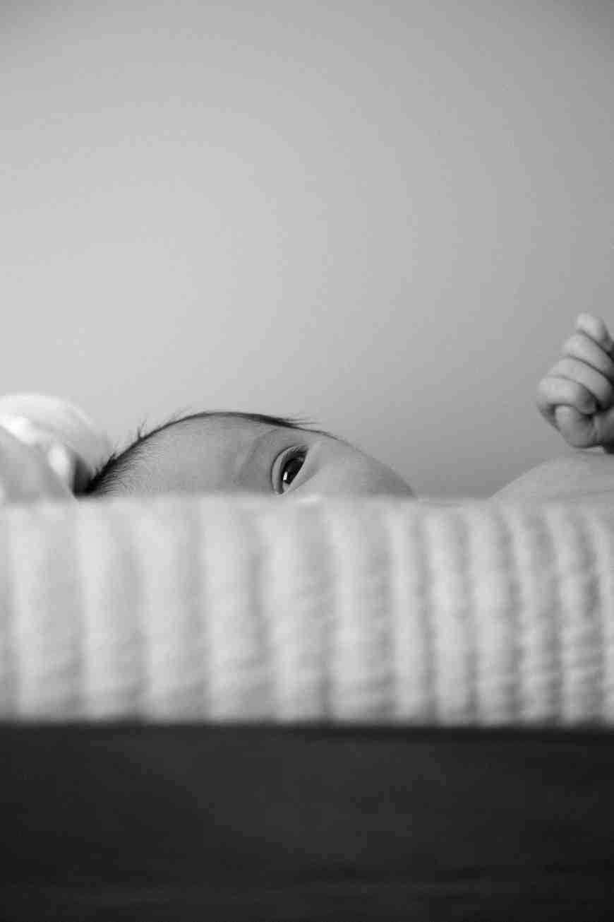 a black and white photo of a striped bed with a baby peaking over the edge. The left side of their face is visible.