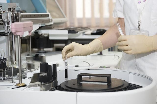 Someone in a laboratory wears gloves on their hands, as they place a test tube filled with liquid into a centrifuge machine. Other laboratory equipment surrounds the room.