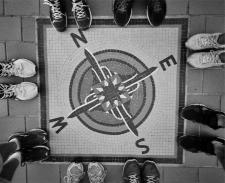 Black and white image of the compass directions--North, East, South and West-- on the ground, with humans wearing shoes surrounding it.