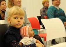 A young blonde white girl, with hair in a pony tail holds a doll. She looks towards the camera but not directly into it.