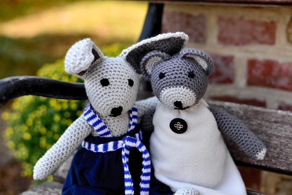 Two crocheted stuffed animals sit on a park bench with arms around each other