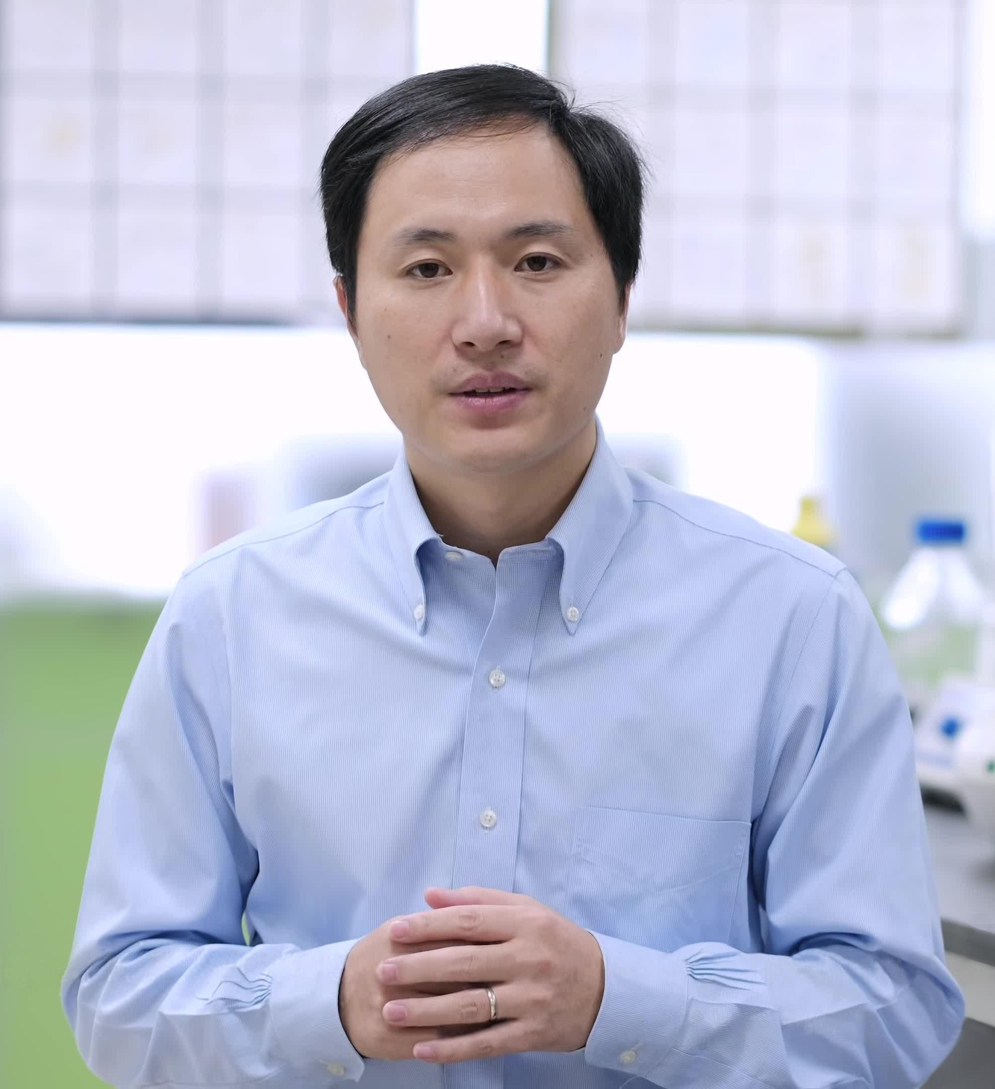 he jiankui in a light blue button up standing in front of a lab. The background is blurred