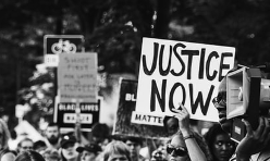 Handwritten sign held up at a protest reads "JUSTICE NOW" in all caps