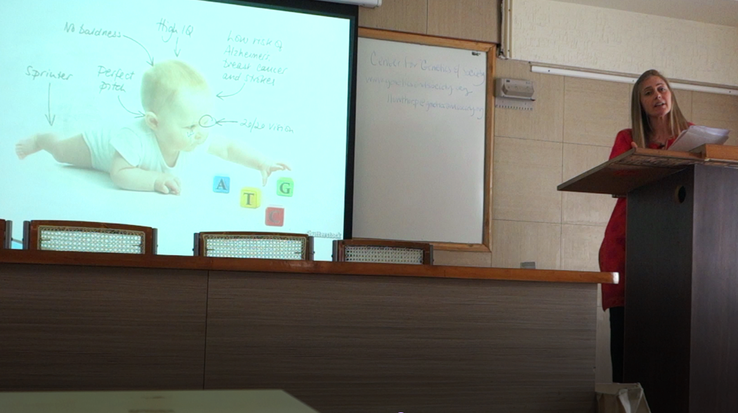 Tall blonde woman in Indian dress giving talk in front of an auditorium with a slide showing a designer baby