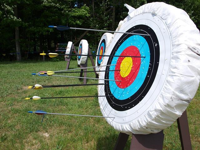 In the foreground, an archery target with several arrows scattered around the target. A line of targets are set up on the grass behind it.