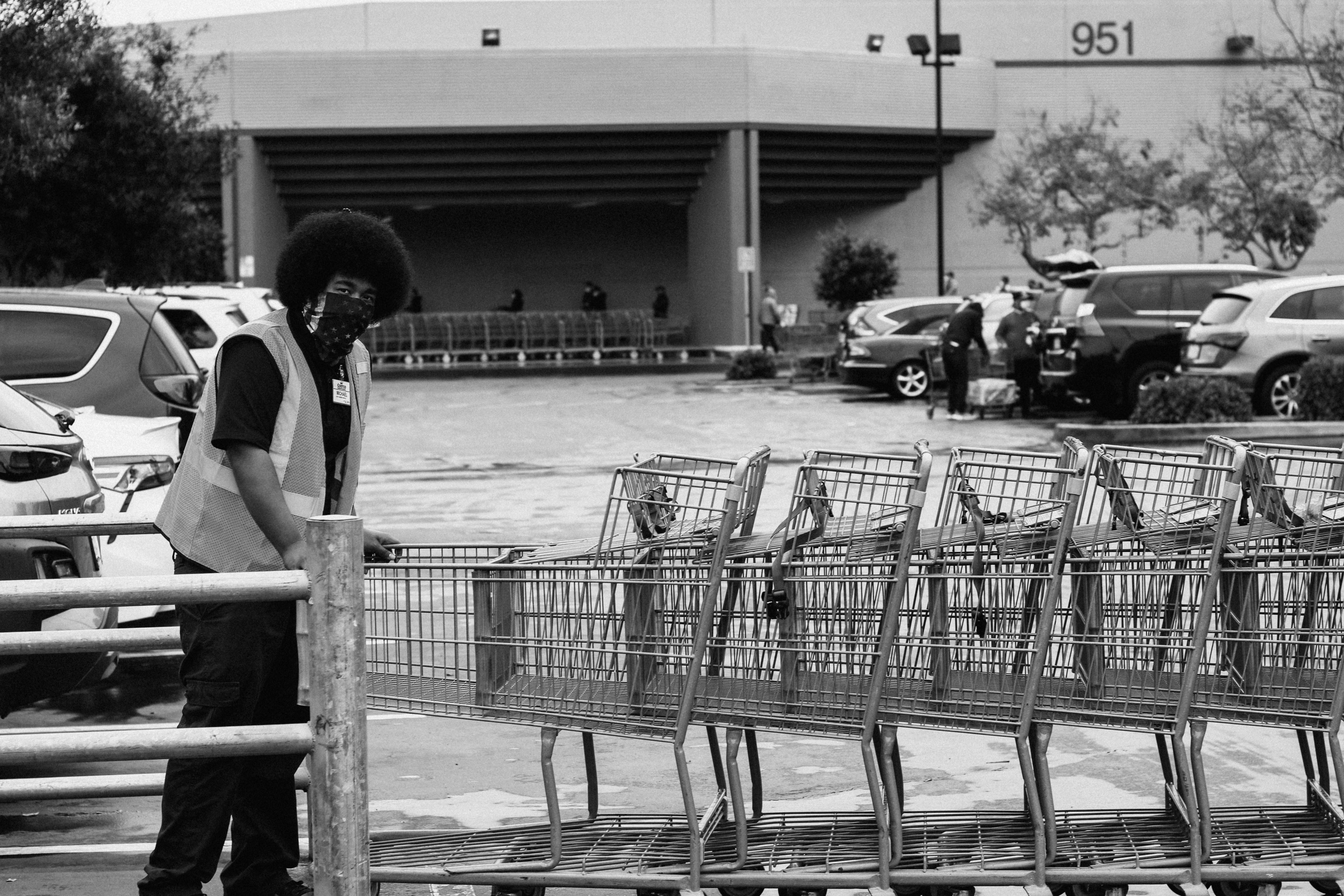 A black man, wearing safety vest and mask made from a bandana, pushes a line of grocery carts in a parking lot