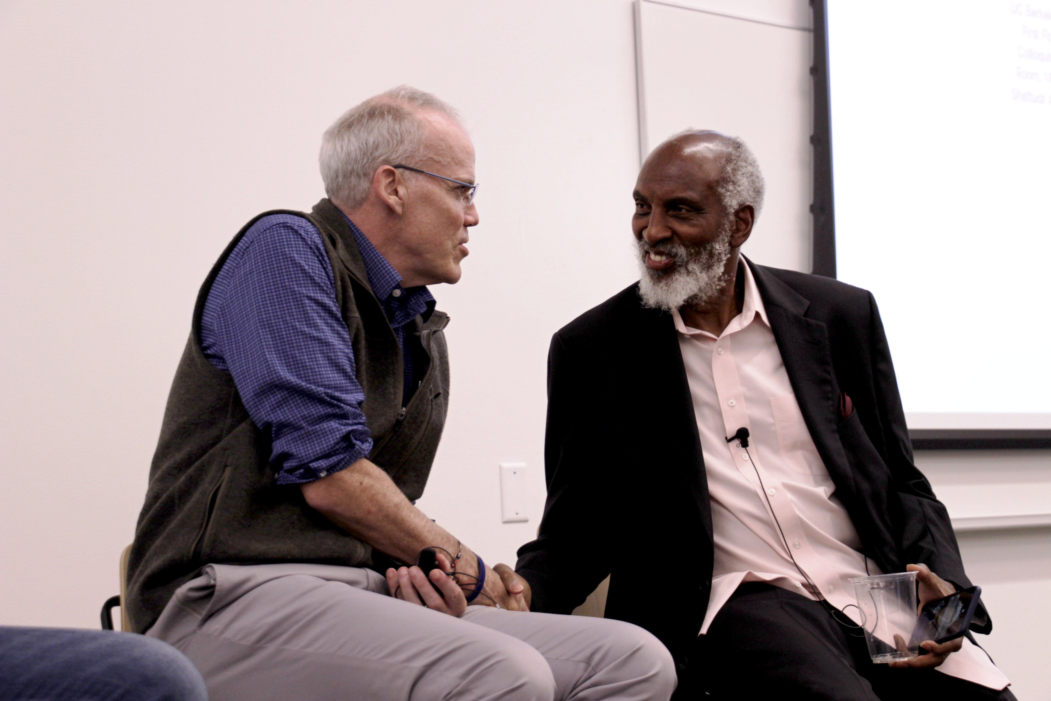 photo of a Bill McKibben, a white man with white hair, and john a. powell, a black man with white hair and beard, shaking hands