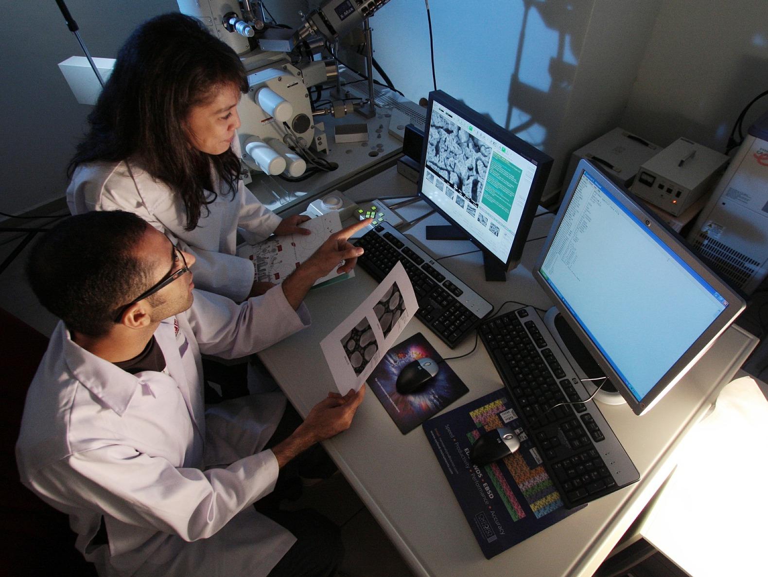 A female and male scientist sitting in a dark lab in front of a screen analyzing images of cells