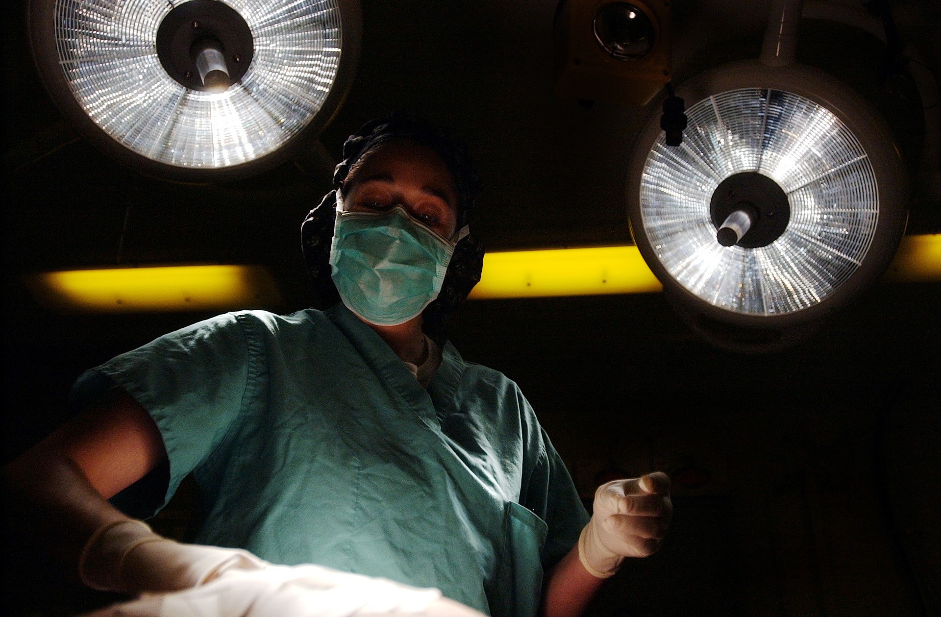 Surrounded by a darkened vignette, a doctor stands looking down towards the camera against two overhead lights. It is as if the camera or viewer is undergoing surgery.