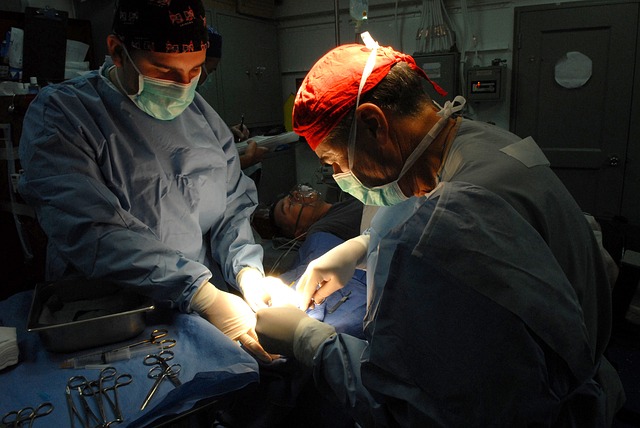 Two surgeons in uniform perform surgery on a patient, with tools in their hands and looking down at the patient.