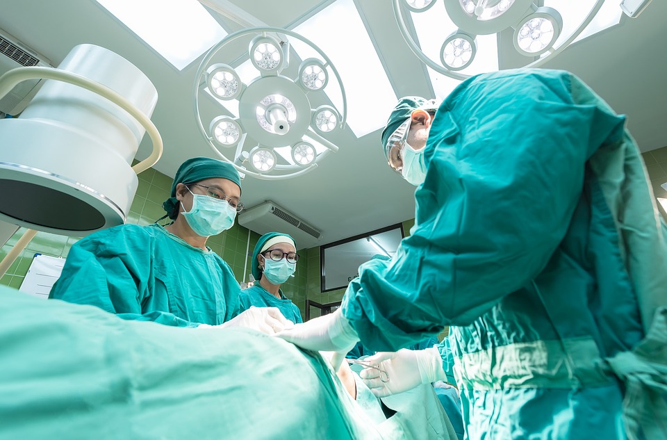 Setting is in bright surgery room. 3 individuals are surround a body covered in a teal sheet and they are all wearing full teal hospital scrubs and white face masks. 
