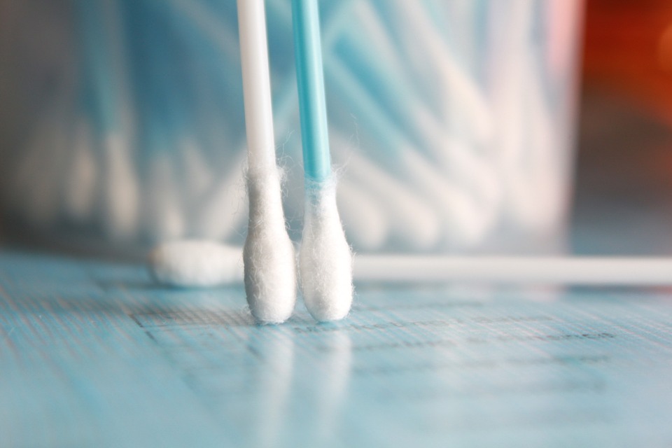 Two Q-tip swabs are vertically positioned next to each other. Blurred in the background, there is a container filled with other q-tips.