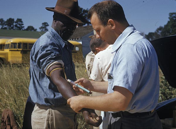 An African Americna male patient looks carefully at a needle injected by a white researcher.