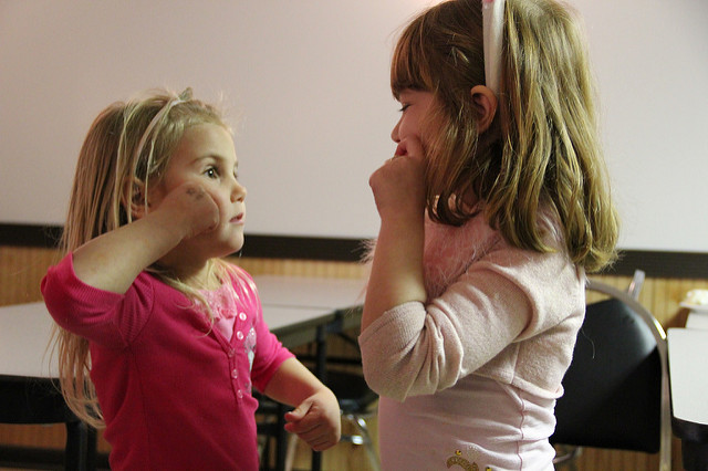 Two young girls face each other, and both appear to be using American Sign Language to communicate. They seem to be signing gum, candy, or apple.