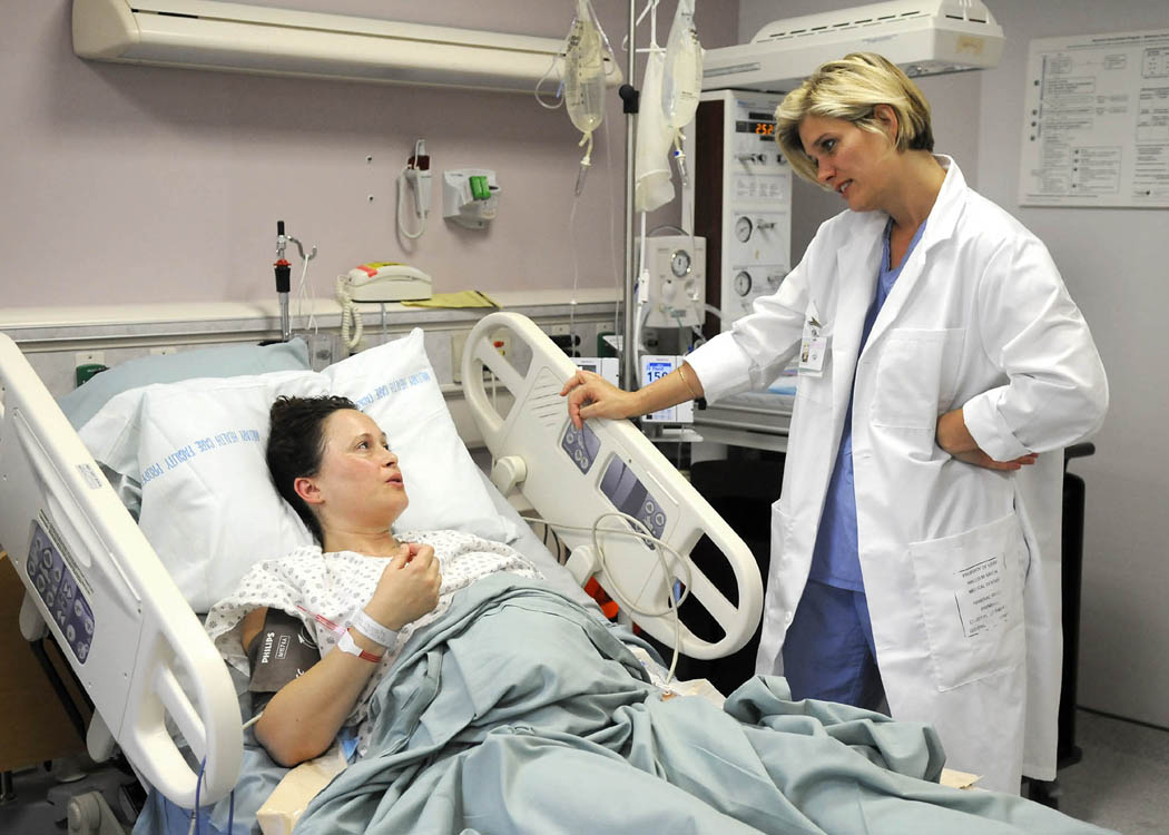 A woman doctor leans over a woman patient's bed, engaged in a conversation The patient has a blood pressure maching attached to her arm.