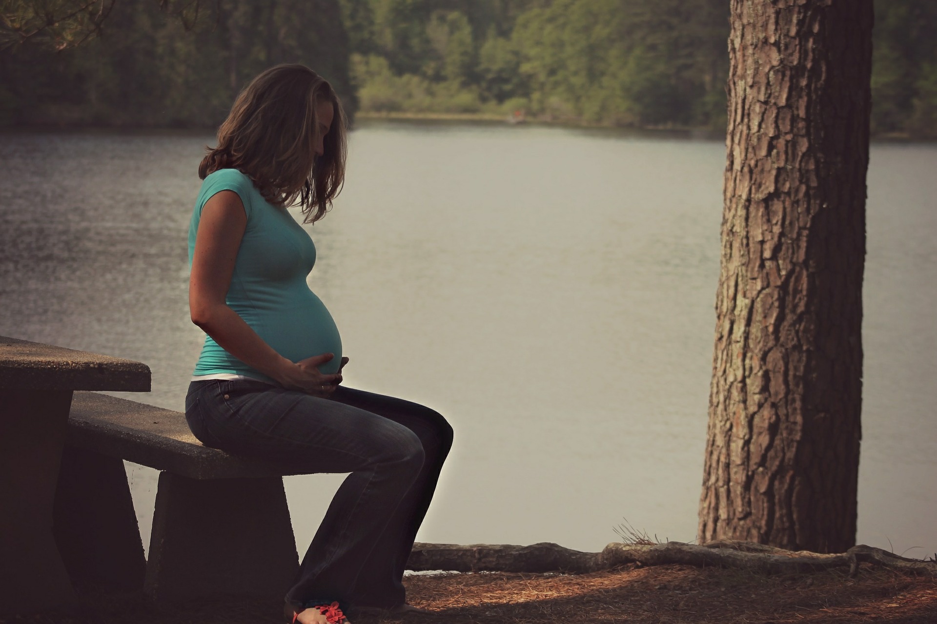 A pregnant woman of color sits at a bench and table, looking down. Behind her, there is a lake and a tree trunk.