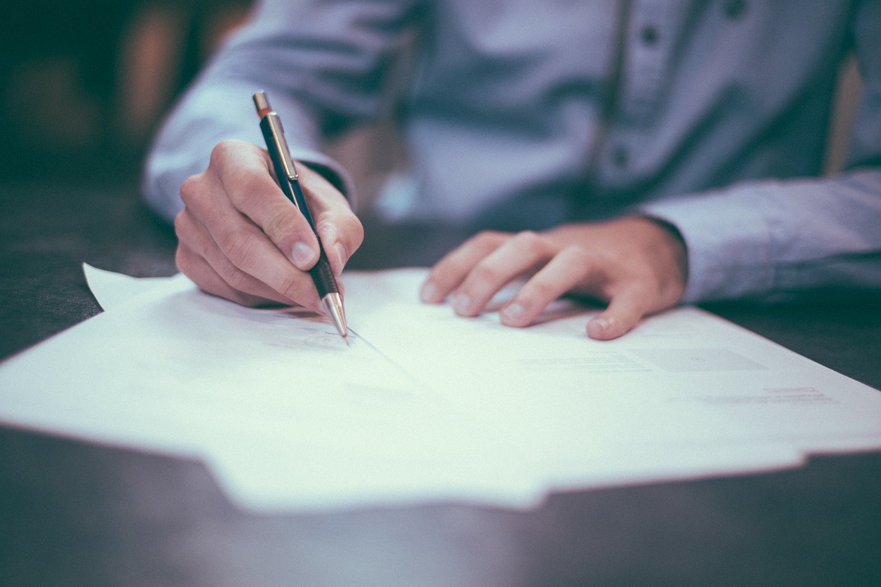 Image of an individual wearing a blue button down shirt signing papers on a dark brown table. 